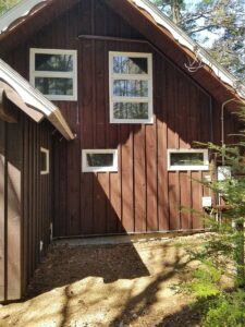 Brown wooden house with white-trimmed rectangular windows, steep roof, and partial shade cast on the exterior wall.