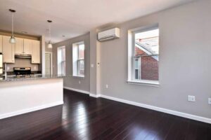 Modern, empty apartment interior featuring dark wood floors, light gray walls, a kitchen with white cabinets and stainless steel appliances, and large windows with natural light.