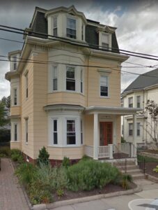 Three-story beige house with a mansard roof, bay windows, and a covered front porch, surrounded by shrubs and a paved walkway.