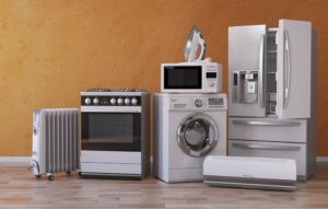 A row of home appliances including a radiator, stove, washing machine, microwave with an iron on top, refrigerator, and air conditioner against an orange wall.
