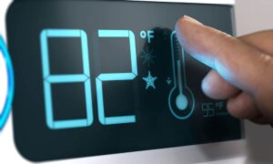 A finger adjusts the temperature setting on a digital thermostat showing 82°F, with icons for heating and cooling displayed on the screen.