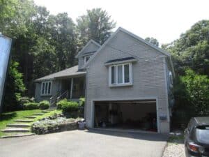 Two-story gray house with attached garage, open garage door showing storage items inside, front steps with greenery, and trees in the background.