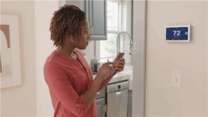 A woman stands in a kitchen using her phone in front of a wall-mounted digital thermostat displaying 72 degrees.
