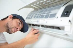 A technician in a blue cap uses a screwdriver to repair or maintain a wall-mounted air conditioning unit.