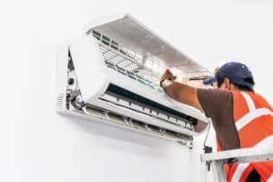 A technician in a safety vest services a wall-mounted air conditioning unit, with the cover open, while standing on a stepladder.