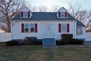 A white, single-story house with gray roof, red shutters, and a central front door, set behind a small lawn and shrubs, with bare trees in the background.