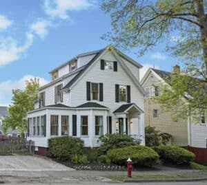 A white three-story house with black shutters, a front porch, and a small garden sits on a residential street next to a yellow house, with a fire hydrant out front.