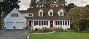 White two-story house with black shutters, red front door, attached double garage, and a well-maintained lawn with shrubs and trees.