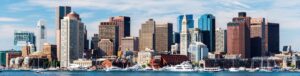 Panoramic view of Boston’s downtown skyline with modern and historic buildings, boats docked along the waterfront, and a partly cloudy sky above.