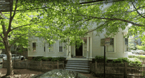 A white wooden house with a front porch and steps is partially shaded by large trees. There is a picket fence and a parking sign in front.