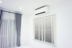 A wall-mounted air conditioner is installed above a window with white blinds in a room with light gray walls and gray curtains.