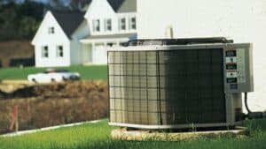 An outdoor air conditioning unit is installed next to the exterior wall of a house, with another house and a car visible in the blurry background.