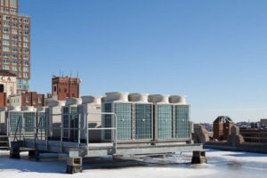 Several large commercial HVAC units are installed on a metal platform on a city rooftop, with buildings visible in the background under a clear sky.