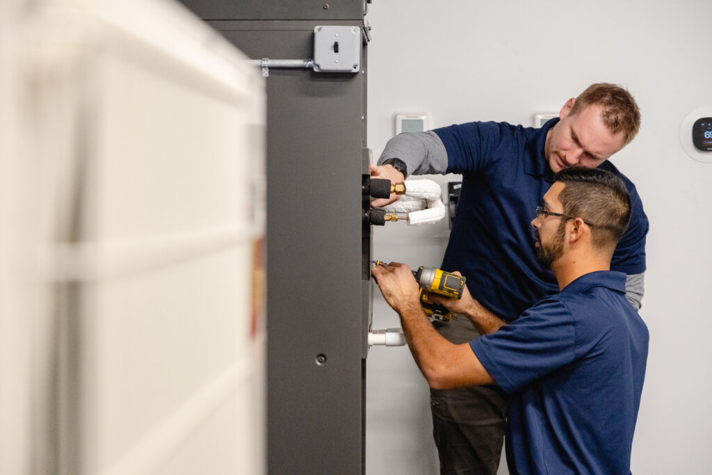 Two HVAC technicians installing or servicing a heating and cooling system inside a mechanical room.