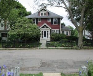 A two-story red house with white trim, surrounded by trees and bushes, sits behind a black wooden fence on a quiet residential street.
