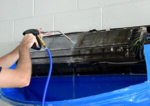 A person uses a high-pressure water spray to clean an air conditioning unit mounted on a wall, with a blue cover below to catch water runoff.