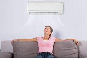 A woman sits on a gray couch, looking up and relaxing under a wall-mounted air conditioner blowing cool air.