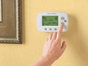 A hand is pressing a button on a digital thermostat mounted on a yellow wall, next to a framed picture.