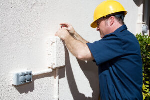 A man in a yellow hard hat and blue shirt uses a screwdriver to work on an outdoor electrical box attached to a light-colored wall.