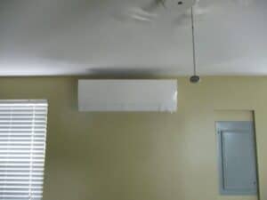 A white wall-mounted air conditioning unit is installed near the ceiling next to a window with blinds and an electrical panel on a beige wall.