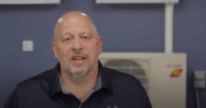 Bald man with a goatee wearing a dark polo shirt sits indoors in front of an air conditioning unit and a blue wall.