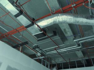 Exposed industrial ceiling with HVAC ductwork, metal pipes, and red conduit lines in an unfinished building interior.