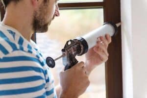 A man in a striped shirt uses a caulking gun to seal the edge of a window frame against a white wall.