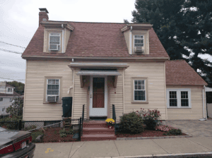 A beige two-story house with a red roof, dormer windows, a small front porch, flower beds, and a parked car in front on a cloudy day.