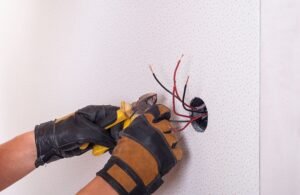 A person wearing protective gloves uses wire cutters on electrical wires protruding from a hole in a wall.