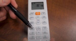 A hand uses a black pen to press a button on a white air conditioner remote control displaying 18°C on a wooden surface.