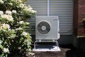 Outdoor air conditioning unit installed next to a house with white siding, near a brick wall and flowering shrubs.