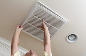 A person removes a ceiling air vent cover, possibly for cleaning or maintenance, in a room with a recessed light nearby.