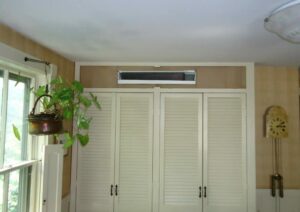 A wall-mounted air vent is installed above cream-colored double closet doors in a room with striped wallpaper and a window with a hanging potted plant.