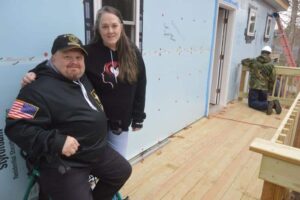 A man and woman pose on a wooden deck of a house under construction, while another person works near the wall in the background.
