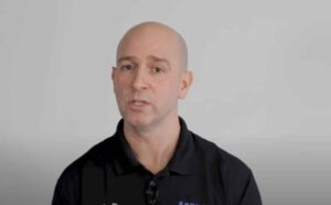 A bald man wearing a black collared shirt speaks while looking slightly past the camera, against a plain light background.