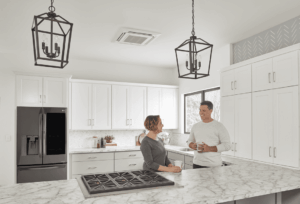 Two people stand and talk in a modern kitchen with white cabinets, marble counters, black appliances, and pendant lights.