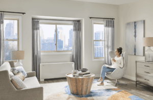A woman sits in a modern living room, holding a mug and looking out large windows at a city skyline. The room is bright, with neutral decor and contemporary furnishings.