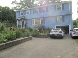 A two-story blue house with white trim, a front garden, and two white cars parked in the driveway on a sunny day.