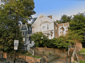 Two large, multi-story houses with gabled roofs and porches sit on a raised lot surrounded by trees and a stone retaining wall, next to a 30 mph speed limit sign.