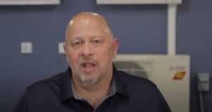 A middle-aged man with a shaved head and short beard speaks while facing the camera, with electrical equipment visible in the background.