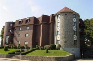 A four-story brick and stone apartment building with cylindrical towers and manicured landscaping, photographed on a sunny day.
