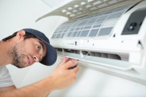 A person in a cap uses a screwdriver to repair or maintain an air conditioner mounted on a wall.