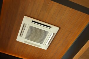 A white ceiling-mounted air conditioning vent is installed on a wooden panel ceiling.