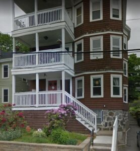 Three-story brown house with white trim, featuring three covered balconies and a front garden with blooming flowers.