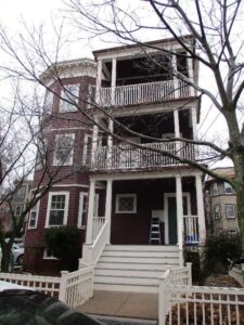 Three-story maroon house with white trim, front porch, balconies on each floor, and leafless trees in front; a ladder is on the porch.