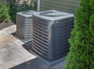 Two outdoor air conditioning units sit on concrete pads next to a house, surrounded by greenery and stone pavers.