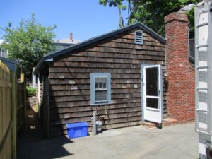A small wooden-sided building with a brick chimney, single window, and a white screen door. A blue recycling bin and utility meters are placed against the wall.