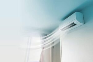 Wall-mounted air conditioner blowing cool air into a bright room with a window covered by sheer curtains.