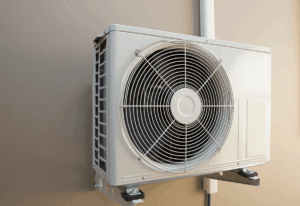 An outdoor air conditioning unit mounted on a beige wall, featuring a large visible fan covered by a metal grille.