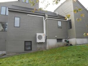 A gray house with vertical and horizontal siding features windows, an outdoor HVAC unit mounted on the wall, a garden hose, and a hose reel on a grassy lawn.